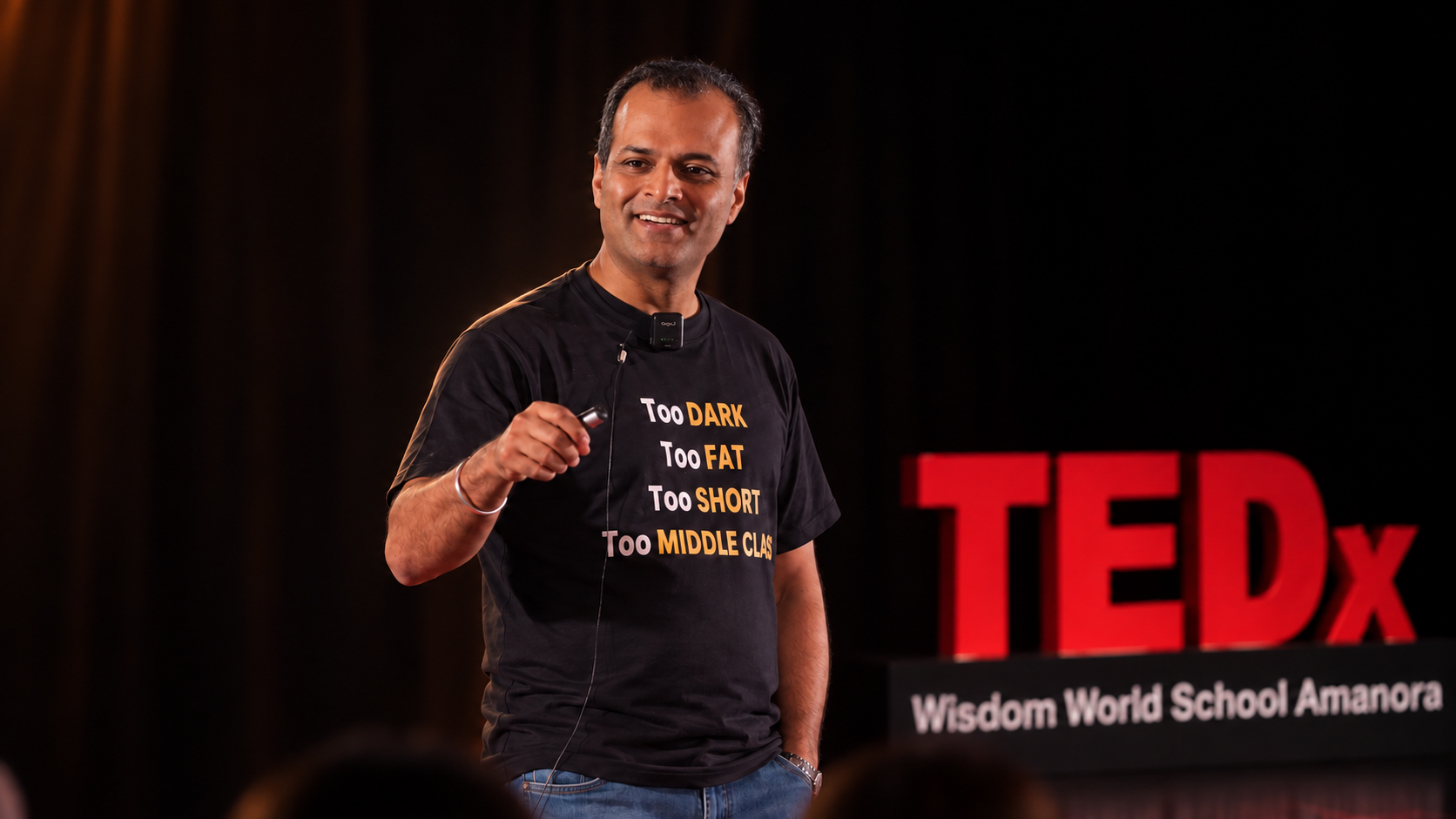Speaker on the TEDx stage, wearing a t-shirt printed with the words Too Dark, Too Fat, Too Short, Too Middle Class