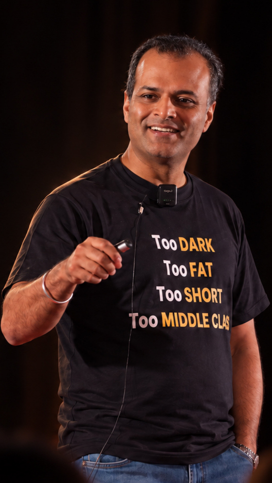 Speaker on the TEDx stage, wearing a t-shirt printed with the words Too Dark, Too Fat, Too Short, Too Middle Class